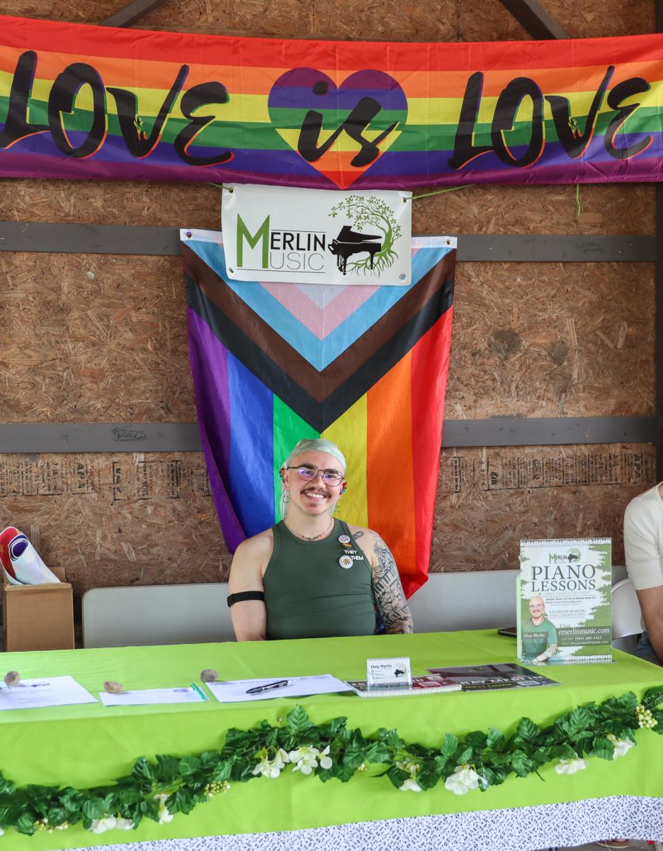 Vendor Merlin Music tabling at Mount Pleasant Pride Festival with a Progress Pride flag and "Love is Love" sign behind them.