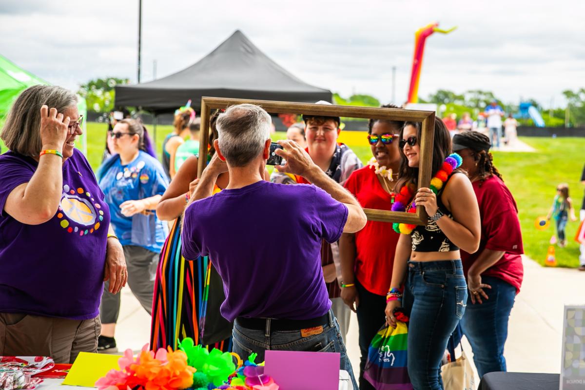 Friends at Great Lakes Bay Pride Festival posing through the opening of an empty picture frame for a photographer.
