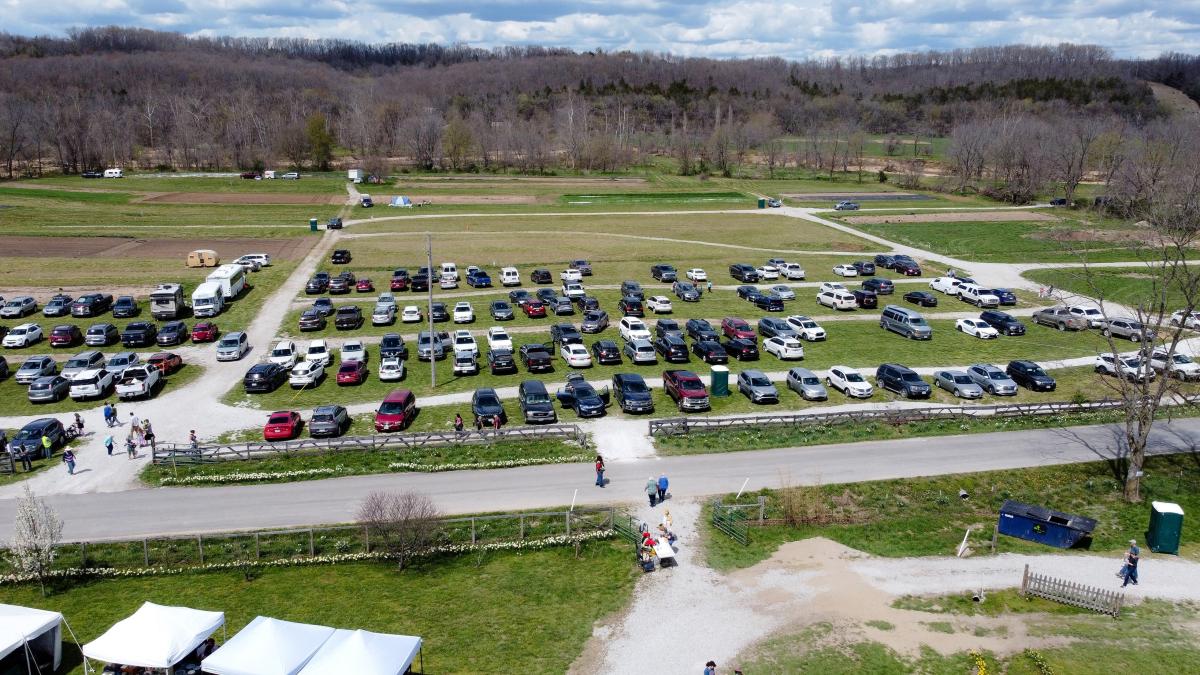 Aerial view of a grass-and-gravel parking lot filled with cars, camping tents in the background, and event tents nearby.