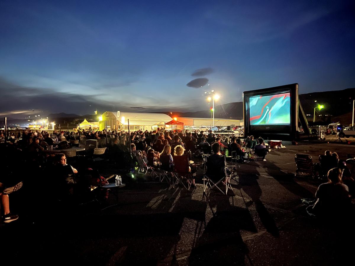 Crowd, at night, sitting in lawn chairs watching a movie on an inflatable screen at Pangborn Airport.