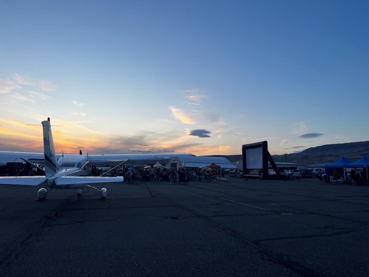Crowd, at dusk, sitting in lawn chairs watching a movie on an inflatable screen at Pangborn Airport.