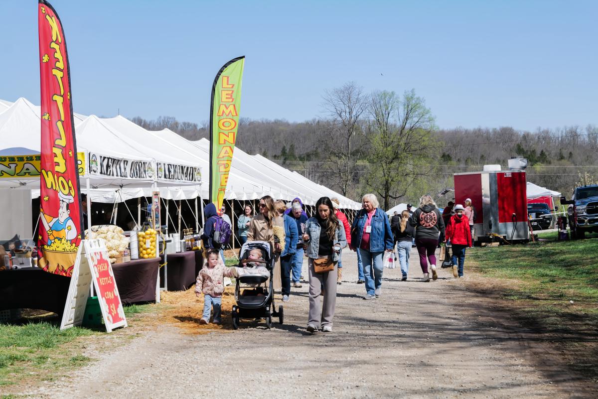 Festival visitors walk a gravel path past kettle corn and lemonade vendors with colorful banners and white tents on a sunny day.